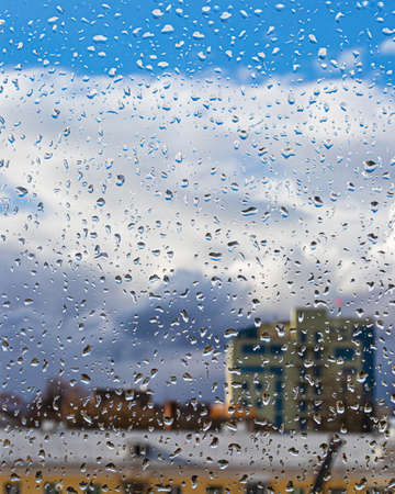Rain drops on window glasses surface with blue sky, buildings and clouds background. The concept of...