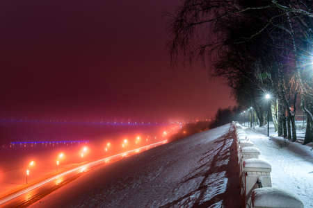 Winter park at night with glowing lanterns, view to road with car motion, pavement and trees in foggy weather.の写真素材