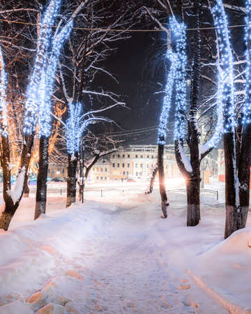 Winter park at night with christmas decorations, glowing lanterns, pavement covered with snow and trees.の写真素材