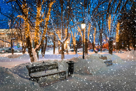 Snowfall in a winter park at night with christmas decorations, glowing lanterns, bench covered with snow and trees.の写真素材
