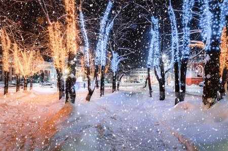 Snowfall in a winter park at night with christmas decorations, glowing lanterns, pavement covered with snow and trees.の写真素材