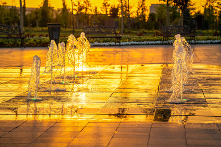 Small fountains on the sidewalk, illuminated by sunlight at sunset or sunrise in the park at summertime.の写真素材