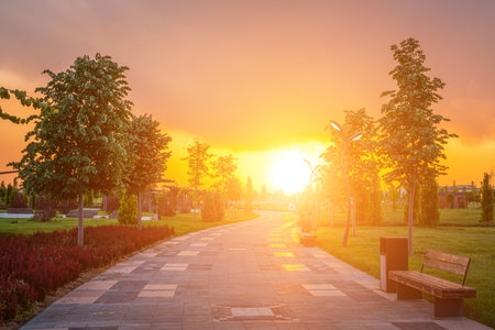 City park in early summer or spring with pavement, lanterns, young green lawn, trees and dramatic cloudy sky on a sunset or sunrise. landscape.の写真素材