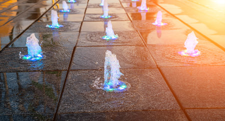 Small fountains on the sidewalk, illuminated by sunlight at sunset or sunrise in the park at summertime.の写真素材