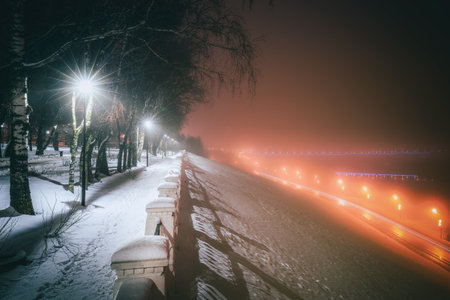 Winter park at night with glowing lanterns, view to road with car motion, pavement and trees in foggy winter weather. vintage film aesthetic.の写真素材