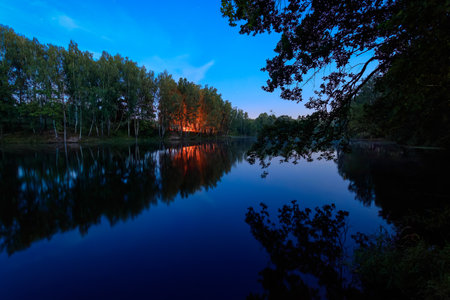 Night landscape with bonfire reflection on water surface, blue sky, forest, trees, river and branches over the water. Nature background for wallpaper or poster idea. Rural scene.の写真素材