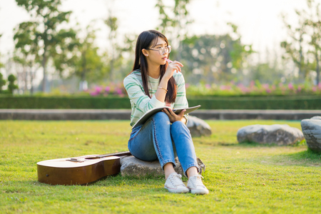 Teenage girl wearing glasses sitting on rock with guitar and books On the football field at sunset.の写真素材