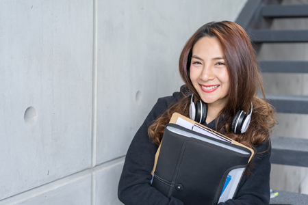 Young student female sit on stairs  with the folder and copy-book and headphone  in campus areaの写真素材