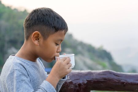 Asian kid boy is drinking water from the glass. With mountain views backgroundの写真素材