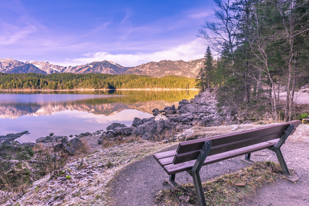 Bench on the shore of an alpine lake - Winter image with a bench located on the shore of the Eibsee lake, from Grainau, Germany. Reflected in its water are the Bavarian Alps mountains.の写真素材