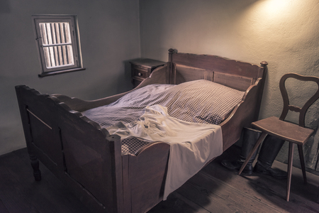 Vintage bedroom with wooden bed and chair - Interior of a rustic bedroom with an aged wooden bed and nightgown on it, a small window, weathered wooden chair and nightstand.のeditorial素材