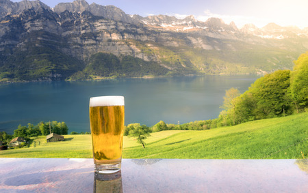 Close-up with a glass of beer on a table with the Alps mountains, green fields and the Walensee lake in the background, on a sunny day of summer.の写真素材