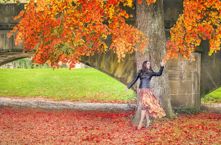 Beautiful young woman dressed in a modern dress and a black jacket, walks under a tree,  reaching...