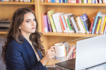 Beautiful, serious businesswoman, in a suit, thinking while having a cup of coffee, at her desk, in...