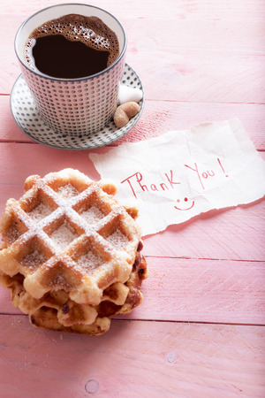 Cute thank you note on a piece of paper with a smiley face on it,  on a pink background, near a hot cup of coffee and fresh waffles, with powdered sugar.の写真素材
