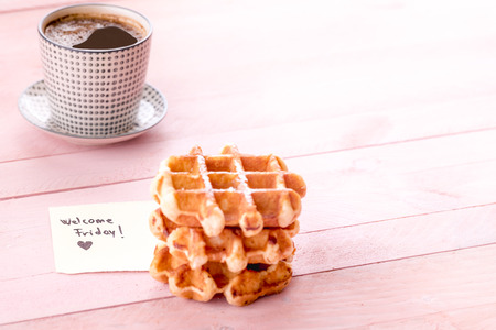 Breakfast with waffles and cup of coffee and a cute note with welcome Friday and a heart, on a pink wooden table.の写真素材