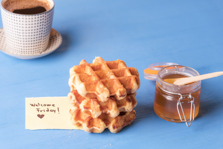 Jar of honey, fresh waffles and hot cup of coffee and a piece of paper with the message welcome Friday and a heart, on a blue wooden background.の写真素材