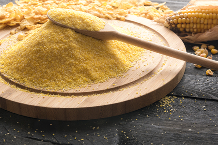Heap of corn flour on a wooden platter with a wooden spoon on top of it, surrounded by corn cob, grains and cereal flakes, on a  black wooden background.の写真素材