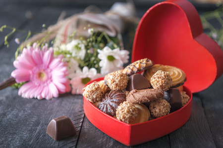 Valentine day theme image with a heart-shaped gift box full of cookies and chocolates, a lovely bouquet of flowers in the background, on a black table.の写真素材