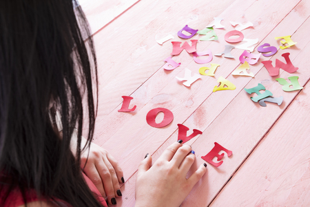 Brunette woman with long hair writing the word love from red paper letters selected from a pile of multicolor letters, on a pink wooden table.の写真素材
