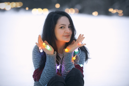 Happy attractive young brunette woman sitting in the snow, holding in her hands colorful lit string lights and playing with them.の写真素材
