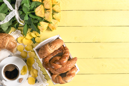 Breakfast table with a cup of coffee, freshly baked croissants and bagels, with flower petals on the yellow wooden table and a big bouquet of roses.の写真素材