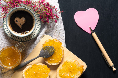 Romantic breakfast with flowers, heart shaped sticky note, bread with jam on a trencher and a cup of coffee with a foam heart, on a black table.の写真素材