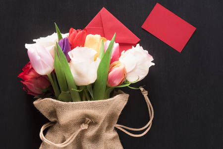 Greeting card with a bouquet of roses and tulips, wrapped in a hessian bag, a closed envelope and a red paper note, on a black wooden background.の写真素材