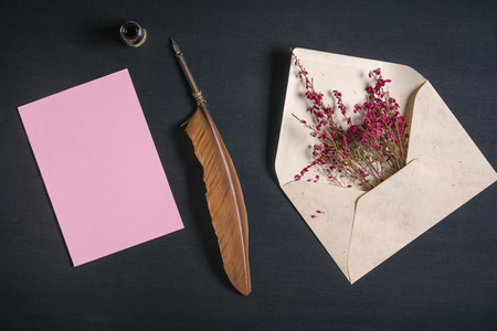 Vintage envelope full with pink flowers, an antique quill pen, an ink pot and a blank message card, on a black wooden background.の写真素材