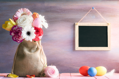Easter banner with a bouquet of flowers in a jute sack, painted eggs and a blank blackboard hanging on a purple wooden background, on a sunny day.の写真素材