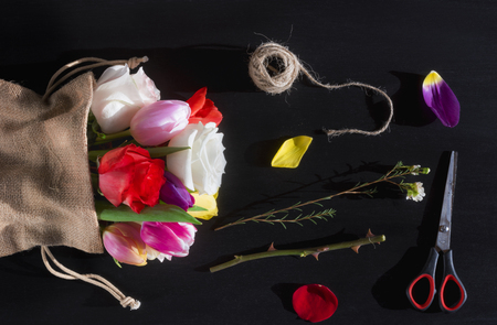 Florist workplace with a bouquet of roses and tulips, wrapped in a jute sack, surrounded by petals, scissors and a clew of linen, on a black table.の写真素材