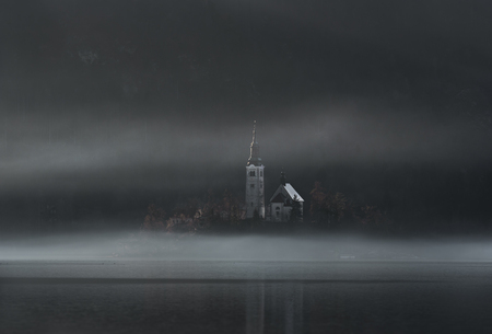 Old church on an island on the Bled lake, surrounded by morning fog and shadow, on a cold day of December, in Bled, Slovenia.の写真素材