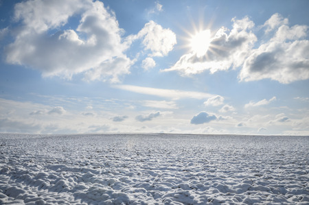 Winter landscape with an empty meadow covered in snow, linked at the horizon with a blue sky and white clouds, under sun rays, in Germany.の写真素材
