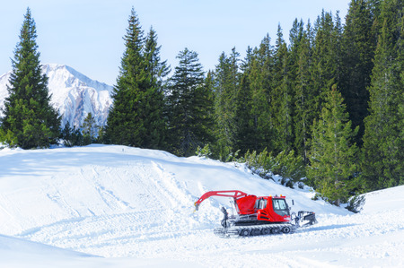 Winter activity in the mountains with a red snow groomer surrounded by snow and green fir forest in...