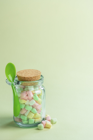 Minimal image with a glass bottle full of various colored tiny marshmallows and a green spoon attached to it, on a greenish paper background.の写真素材