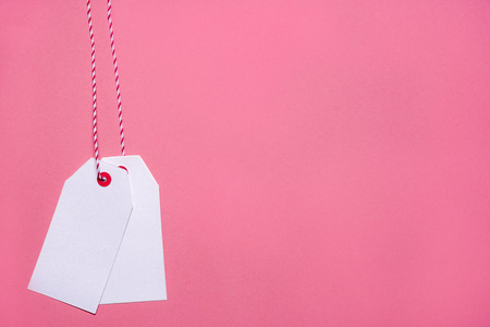Minimalist style image with two blank labels hanging from a red and white string, on a pink paper...
