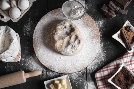 Christmas cookies dough on a wooden platter, surrounded by flour, eggs, cinnamon, chocolate, butter, and a rolling pin, on a rustic black table.の写真素材