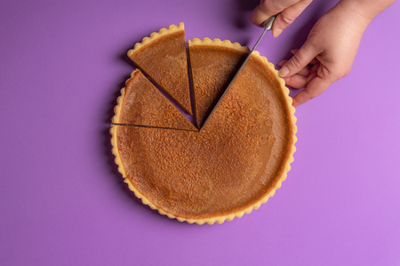 Woman hands slicing a pumpkin pie on a purple background. Minimalist culinary concept. Homemade sweet pie. Traditional holidays dessert. Top view.の写真素材