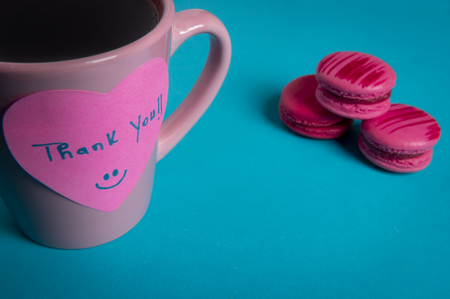 Pink coffee mug and macaroons on a blue background. Paper note with a thank you message fixed on the coffee cupの写真素材