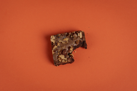 Bitten brownie cake piece on an orange colored background above view in minimalist style. Chocolate dessert decorated with plenty of walnutsの写真素材