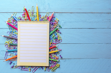 Blank page spiral notebook on a pile of office supplies, colored paper clamps, push pins and wooden...