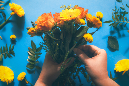 Woman hands making a flower bouquet from vibrant yellow and orange flowers over a blue paper background. Flat lay.の写真素材