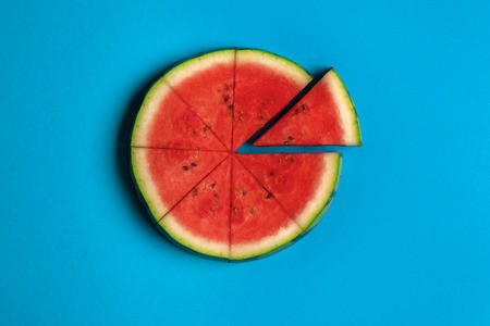 Round slice of watermelon cut in portions with one slice separated, on a blue paper background. Minimalist summer fruit image. Flat lay.の写真素材