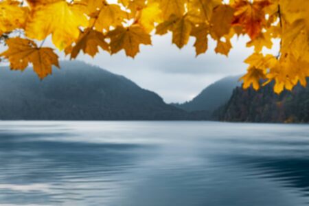 Blurred autumn nature background with yellow leaves as a frame for mountains and blue water lake,...