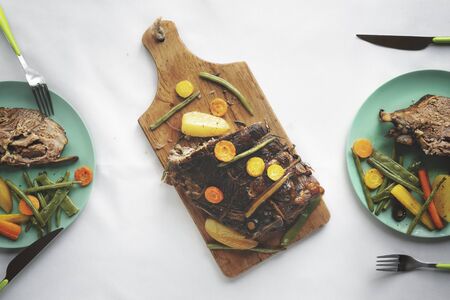 Roasted pork on wooden cutting board and green plates with meat portions and cooked vegetables, on a table with a white tablecloth. Flat lay.の写真素材
