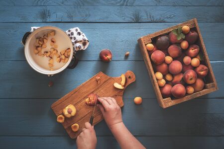 Woman hands cutting into pieces peaches and apricots from a vintage wooden box, on a blue kitchen table. Above view with the making of peaches jam.の写真素材