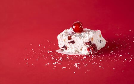 Slice of white Christmas cake with coconut, dried fruit, and cherries, on a red background. Australian traditional sweet food. Winter holiday dessertの写真素材