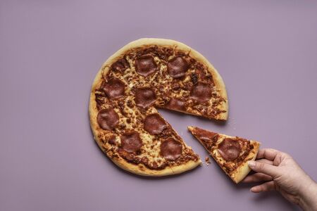Taking one pizza slice. Woman hand grabbing a piece of pizza salami on a purple background. Above view of pepperoni pizza. Traditional italian dinnerの写真素材