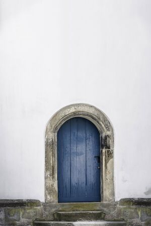 Medieval arched door. Aged wooden door in blue color with stone stairs and white wall. Just one door and an empty white house wall. Old blue entrance.の写真素材