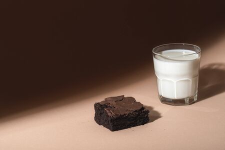 Chocolate brownie cake and a glass of warm milk on a brown background, in sunlight. Indulgence food. Chocolate dessert and milk. Unhealthy breakfast.の写真素材